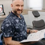 Smiling dentist holding a book, sitting in a dental office with equipment around.